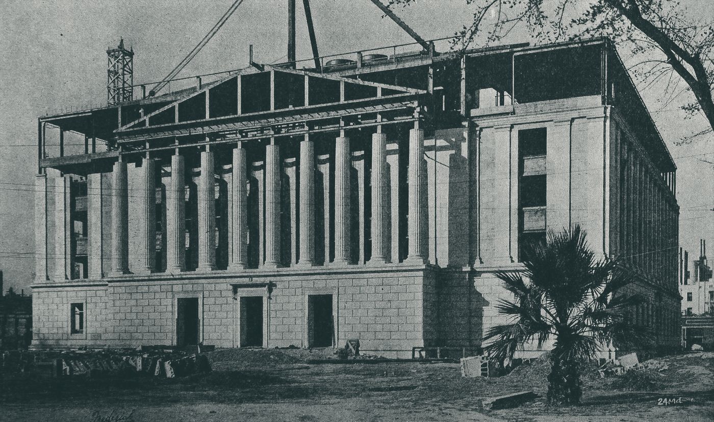 A colonnaded state office building under construction, with steel framing and a crane above the portico
